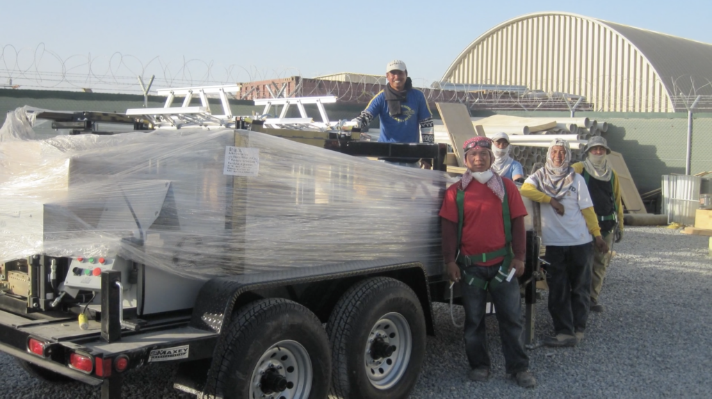 A group of men standing around a rollformer on a trailer, their heads covered to protect them from the heat.