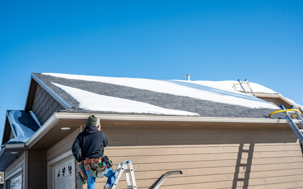 A man standing on a ladder, installing a gutter with snow on the roof.